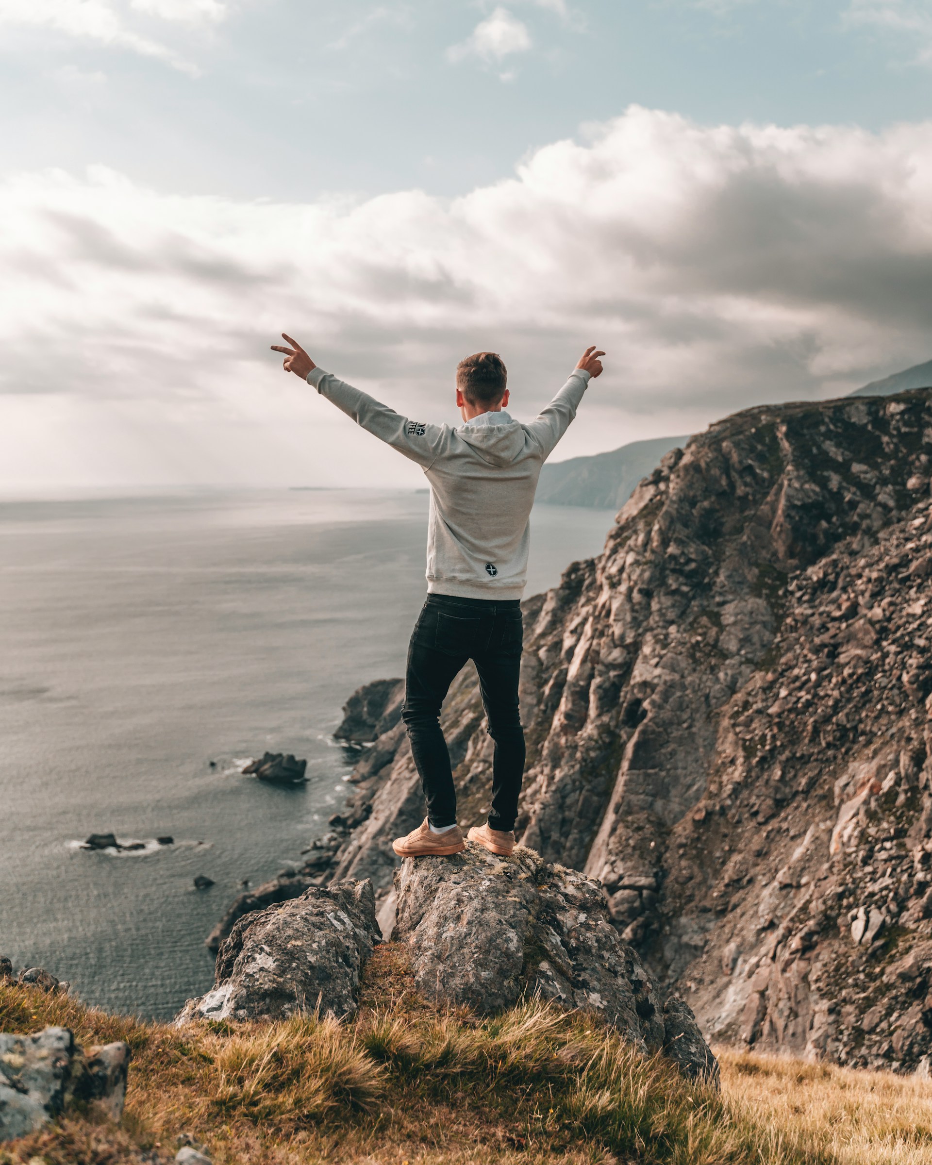 Mies seisoo kädet kohotettuina kallion huipulla meren äärellä. Kuva: Majestic Lukas / Unsplash