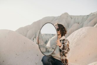 Mies aavikon dyyneillä polvillaan iso pyöreä peili polvella tuettuna- Kuva: Ali Ramazan Ciftci/Unsplash