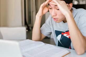 Nuori opiskelee pöydän ääressä avoimen oppikirjan ja kannettavan tietokoneen kanssa. Kuva: RDNE Stock project / Pexels