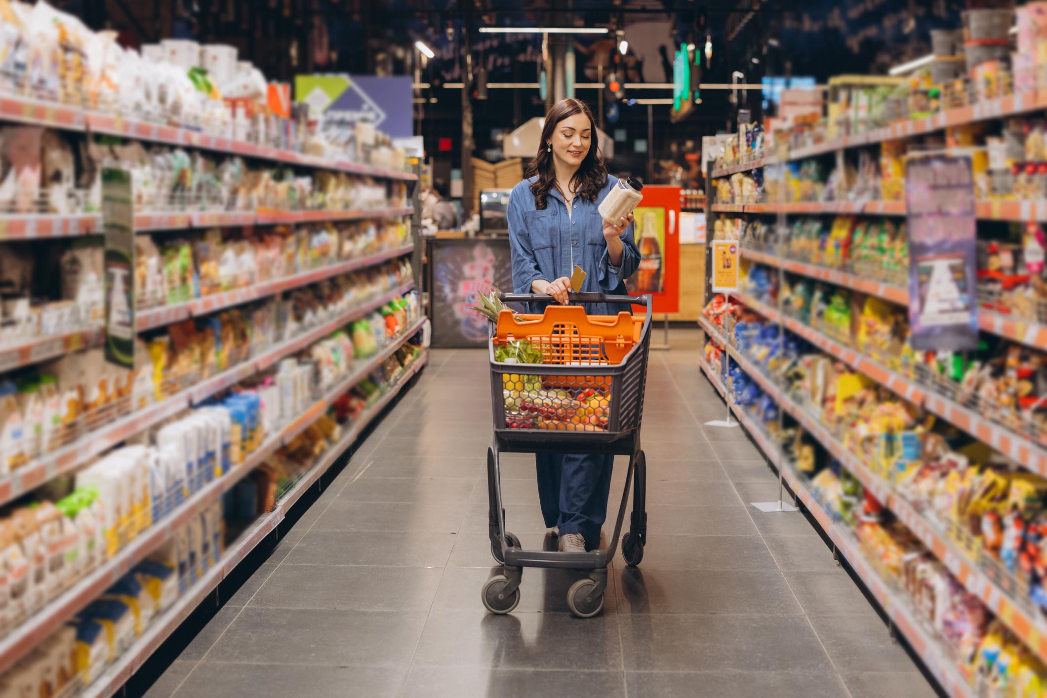 Nainen tarkastelee tuotetta ostoskärryjen kanssa supermarketin pitkällä tuotekäytävällä.