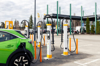 Vihreä sähköauto latauksessa ABC-latausasemalla. Kuvassa näkyy useita sähköautojen latauspylväitä, latauskaapeleita ja ABC-aseman tunnus. Kuva: Osuuskauppa Suur-Savo / STT Info