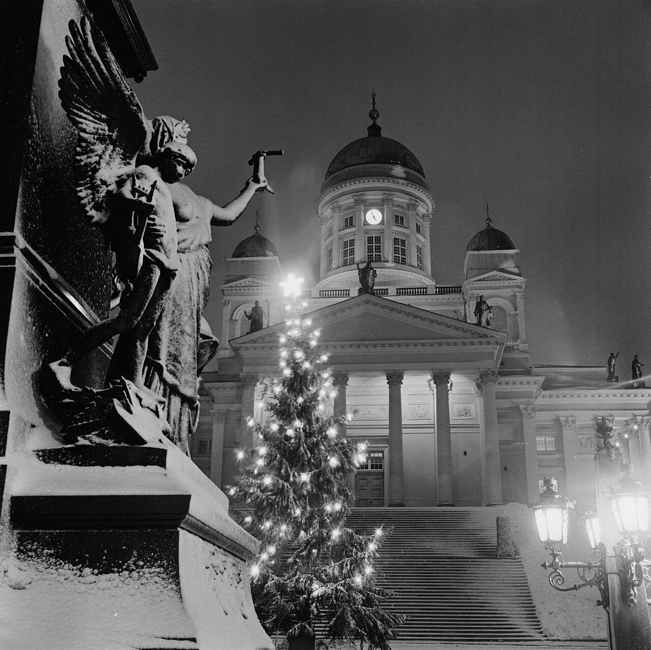 Historiallinen mustavalkokuva Helsingin Senaatintorilta. Valaistu joulukuusi seisoo lumisten portaiden edessä, taustalla Helsingin tuomiokirkko ja vasemmalla suuri patsas.