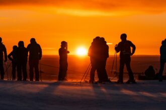 Laskettelijoita ja suksien kanssa seisovia ihmisiä lumisella tunturilla Levillä. Taustalla hehkuu voimakkaan oranssi auringonlasku.
