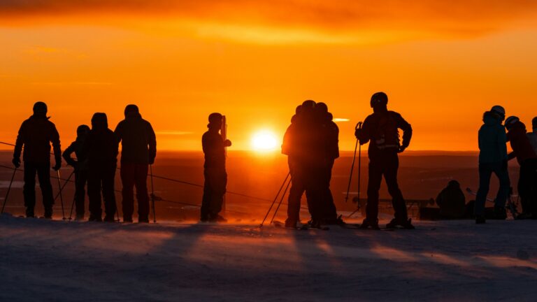 Laskettelijoita ja suksien kanssa seisovia ihmisiä lumisella tunturilla Levillä. Taustalla hehkuu voimakkaan oranssi auringonlasku.