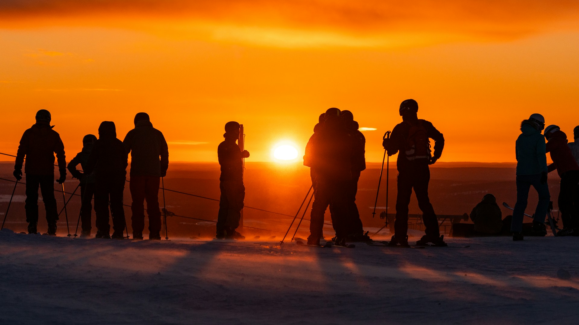 Laskettelijoita ja suksien kanssa seisovia ihmisiä lumisella tunturilla Levillä. Taustalla hehkuu voimakkaan oranssi auringonlasku.