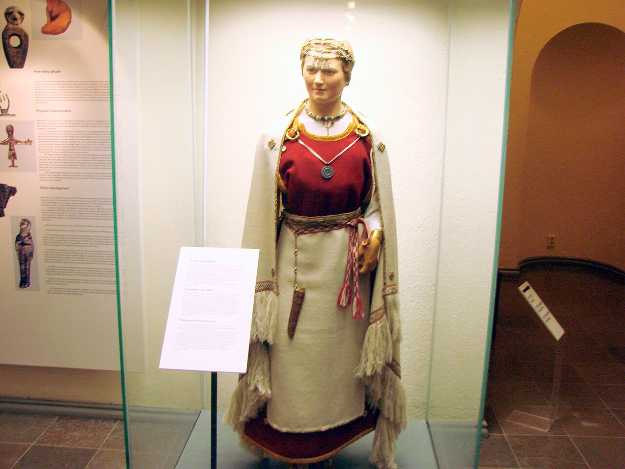 Museossa esillä oleva 1100-luvun suomalaisen naisen muinaispuvun ennallistus, jossa on punainen mekko, vaalea esiliina, viitta ja koristeellisia koruja